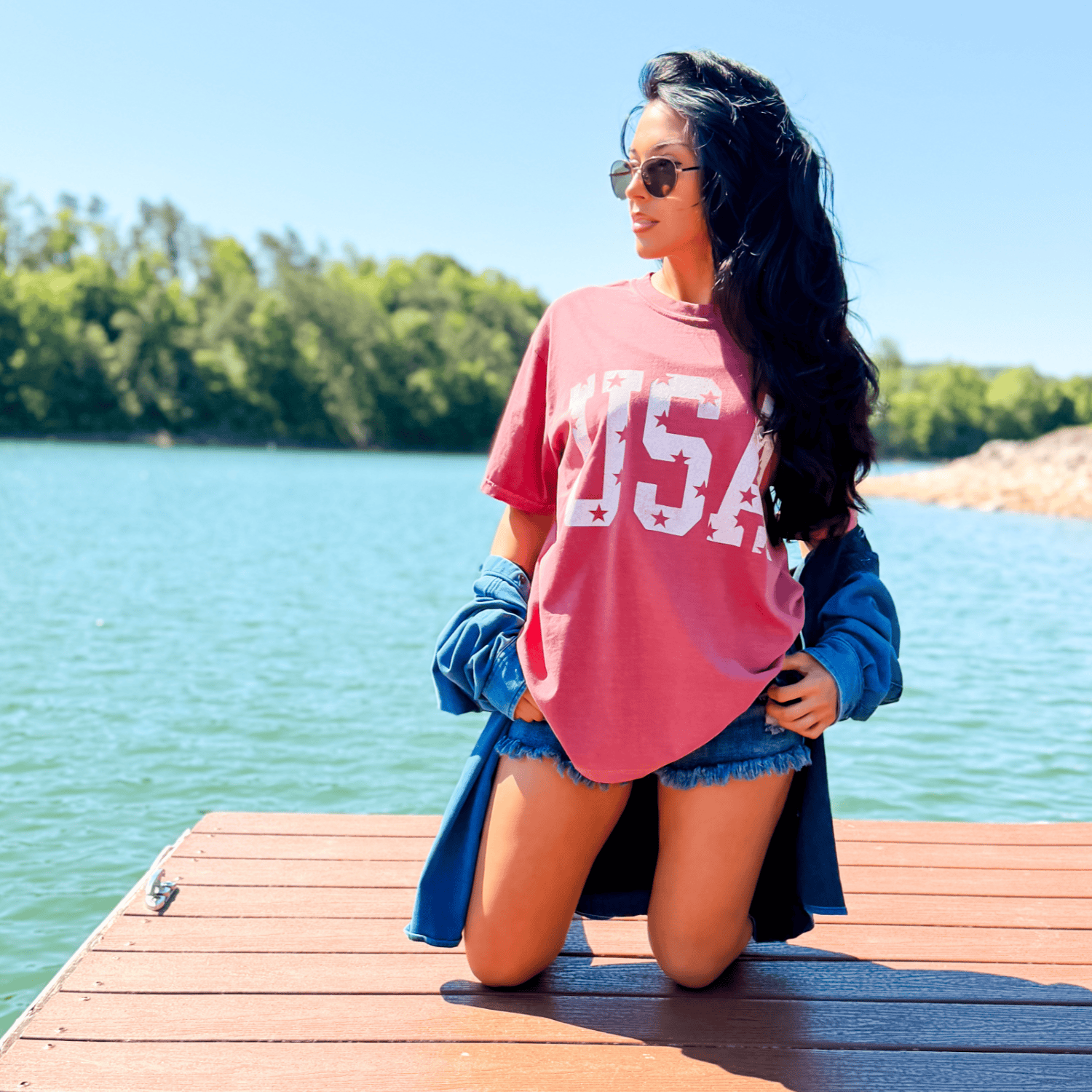 Distressed USA tee in brick red with white stars, worn by a model on a dock by a lake, styled with denim shorts.