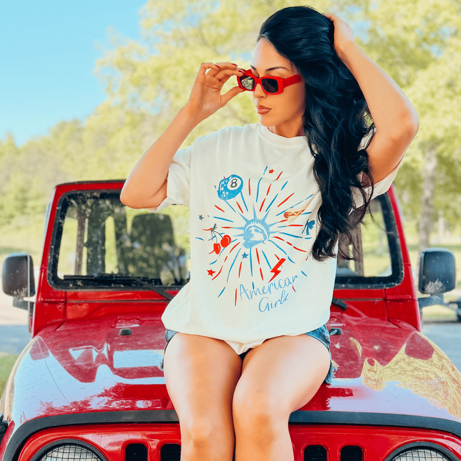 American Girlz tee featuring a colorful graphic design, worn by a woman with long hair, sitting on a red jeep.