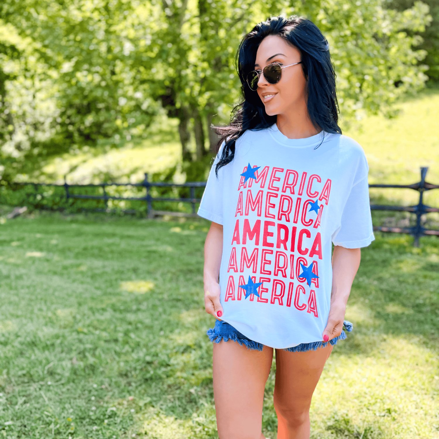 Starry America tee in white with red and blue text, worn by a woman outdoors with sunglasses and denim shorts.