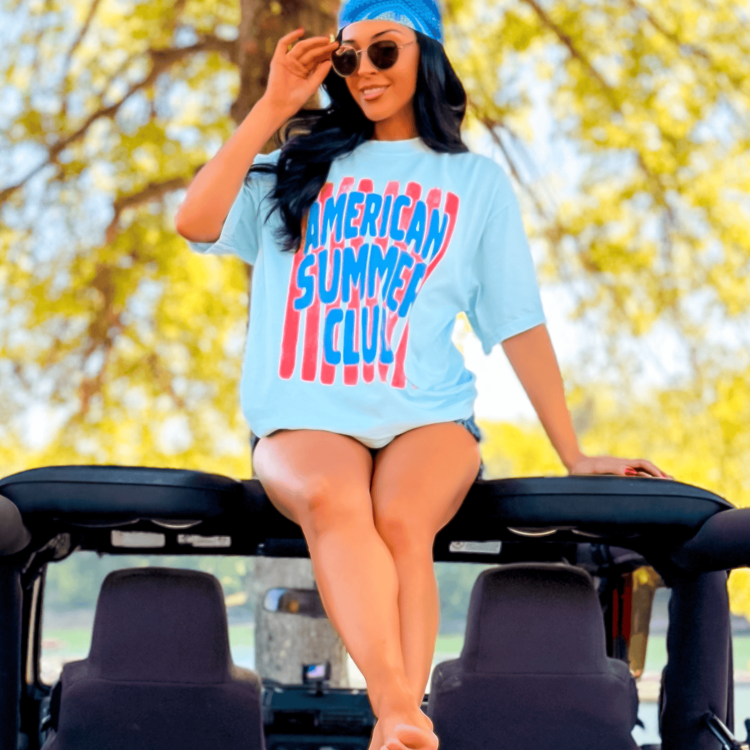 Light blue American Summer Club tee with red text, worn by a woman sitting on a vehicle in a sunny outdoor setting.