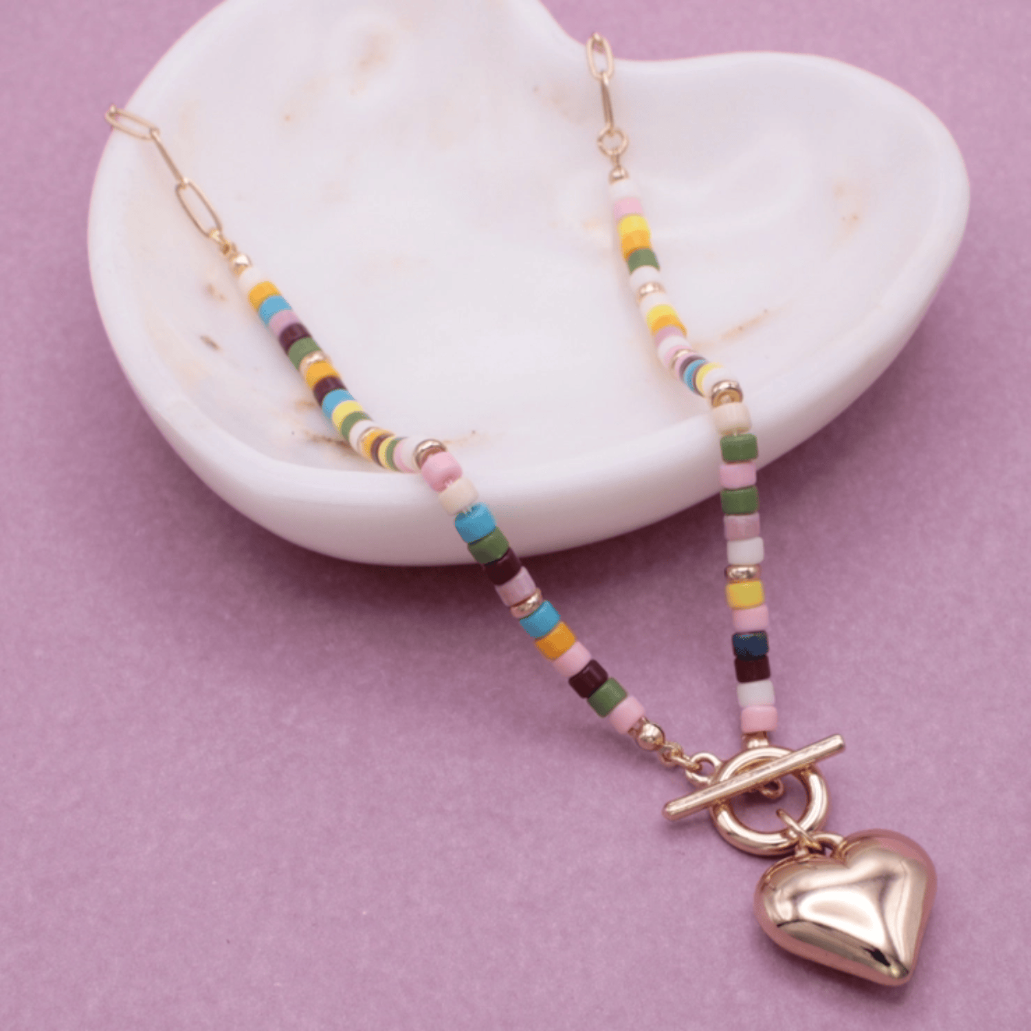 Multicolor gold heart toggle necklace with colorful beads and a shiny heart pendant on a light purple background.