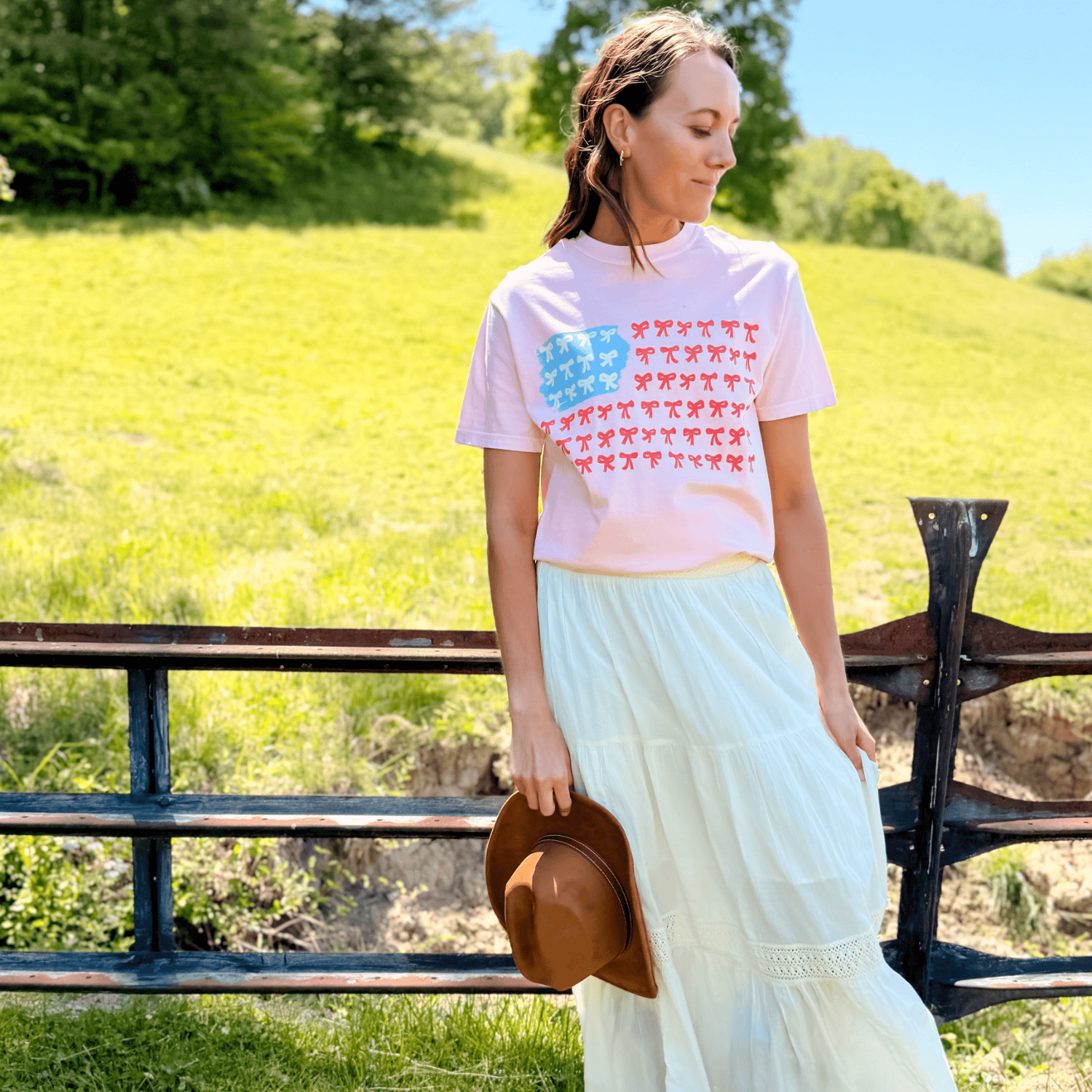 American Flag Bows Tee in pink with blue and red bow patterns, worn by a woman standing near a wooden fence.