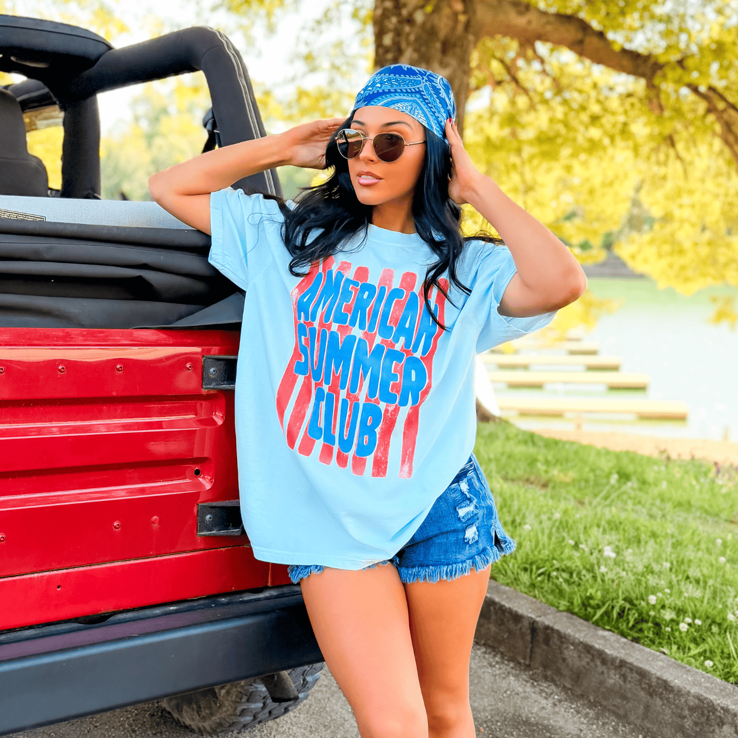 Light blue American Summer Club tee with red graphic, worn by a woman with sunglasses and a blue bandana, near a red vehicle.