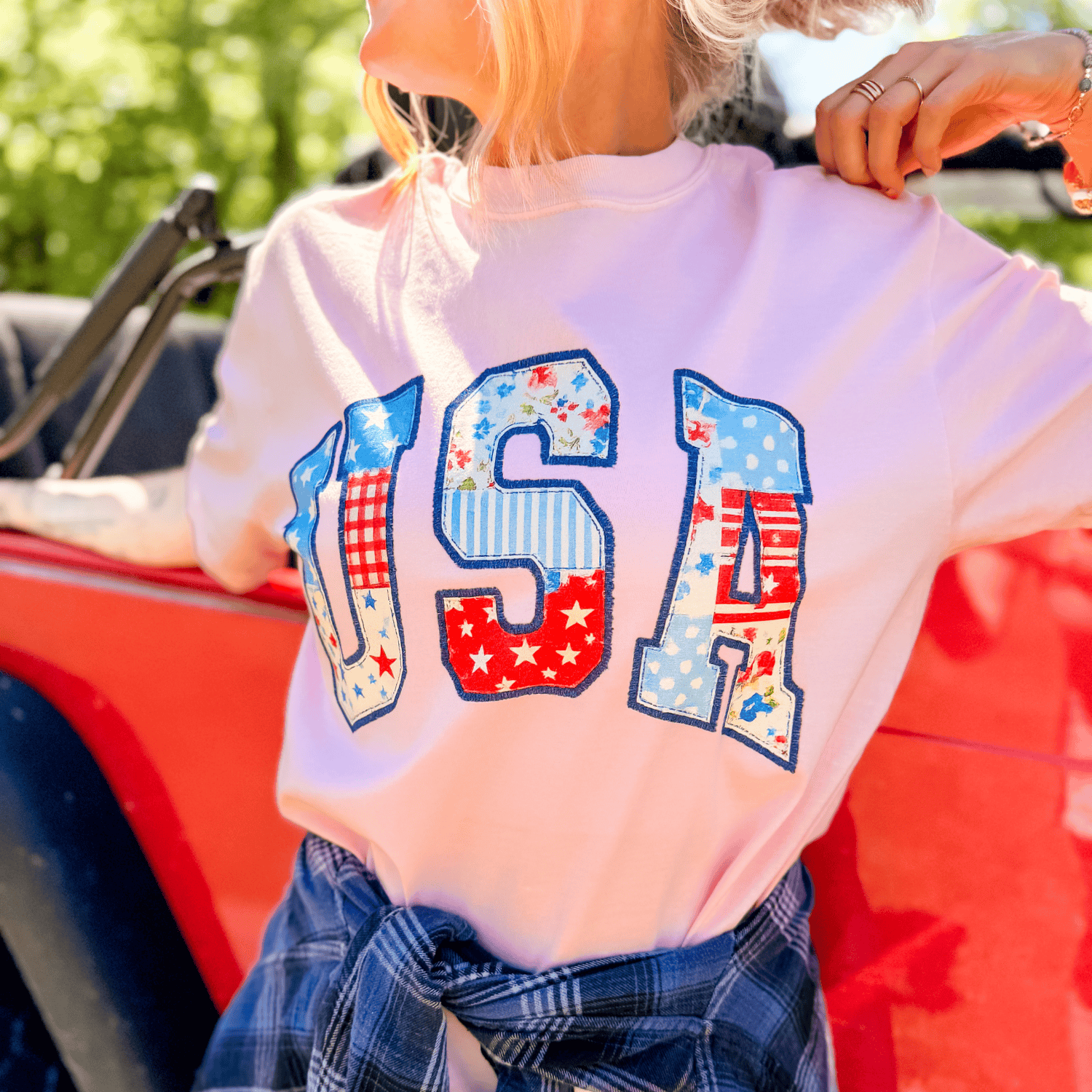 White patchwork USA tee with colorful star and checkered designs, worn by a person near a red vehicle.