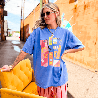 Blue Book Club Tee featuring colorful graphic designs, worn by a woman seated on a yellow chair in an urban setting.