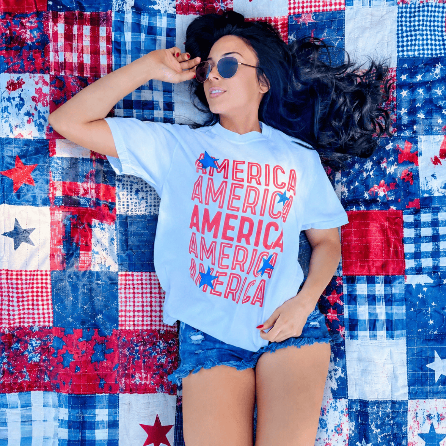 Starry America tee in light blue with red text, worn by a woman lying on a patchwork red, white, and blue quilt.