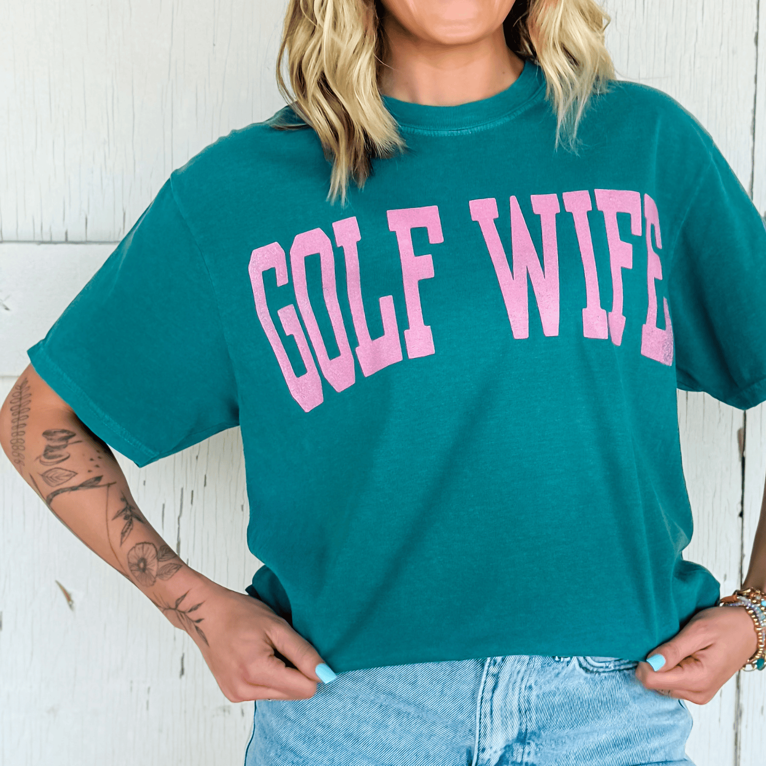 Person wearing a teal t-shirt with 'GOLF WIFE' in pink text against a white wooden background