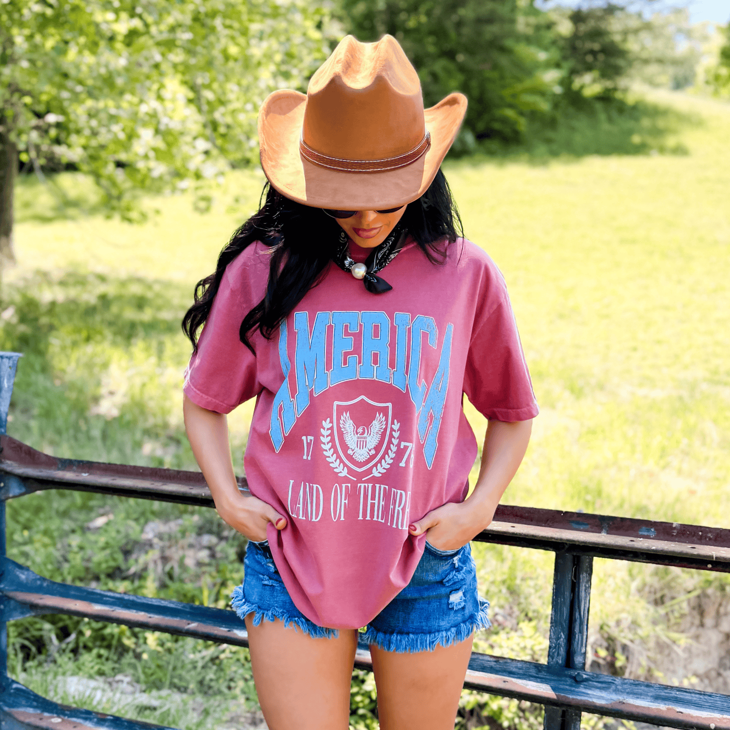 Land of the Free collegiate tee in maroon with blue lettering, worn by a woman in denim shorts and a cowboy hat outdoors.