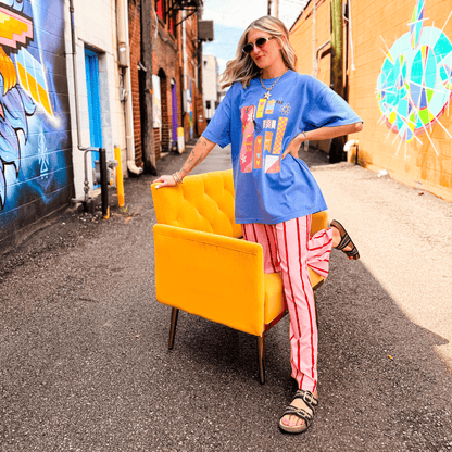 Blue Book Club Tee featuring colorful graphic design, paired with striped pants, posed beside a yellow chair in an urban s...