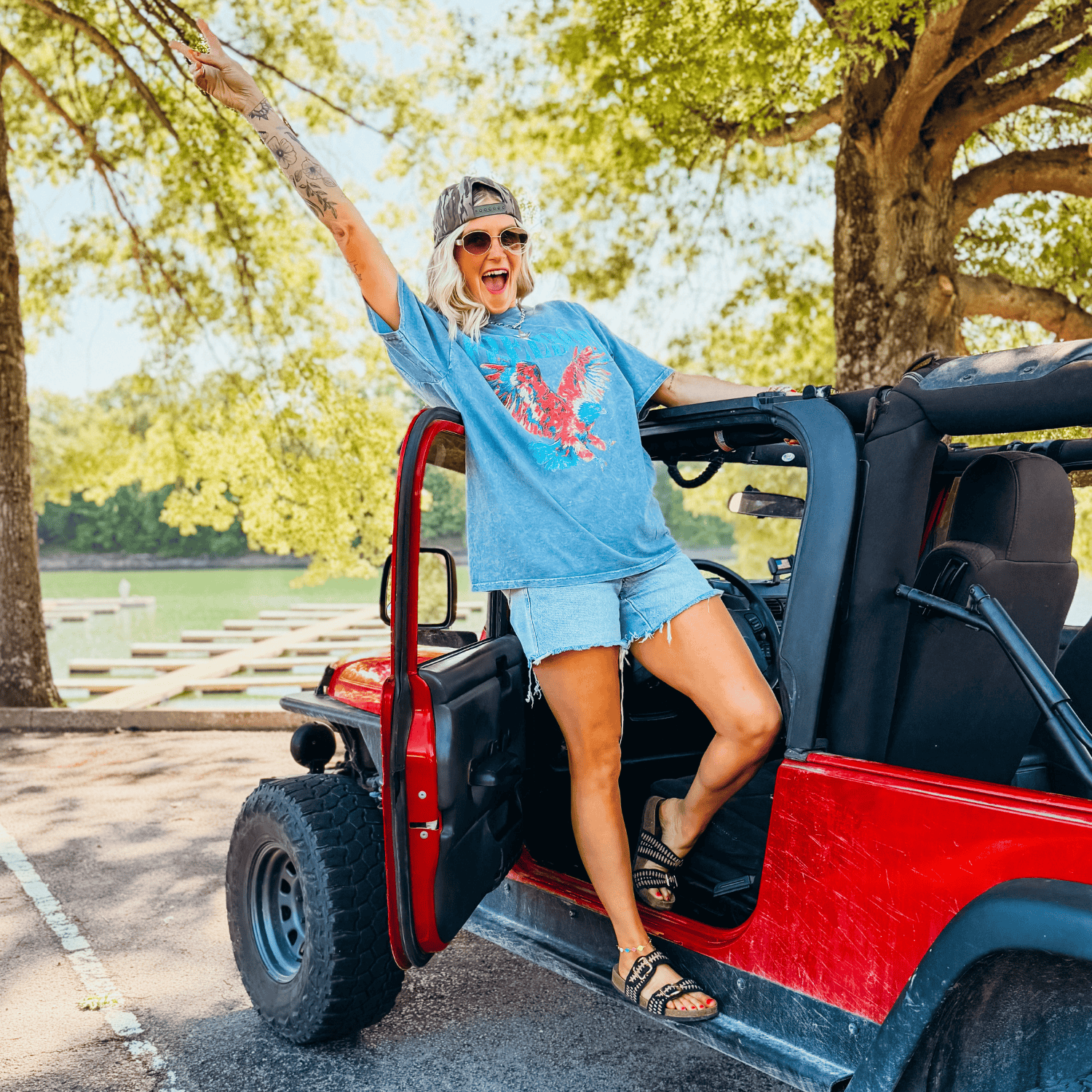 Freedom Mineral Wash Tee in blue, worn by a woman with sunglasses and a cap, standing in a red jeep by a lake.