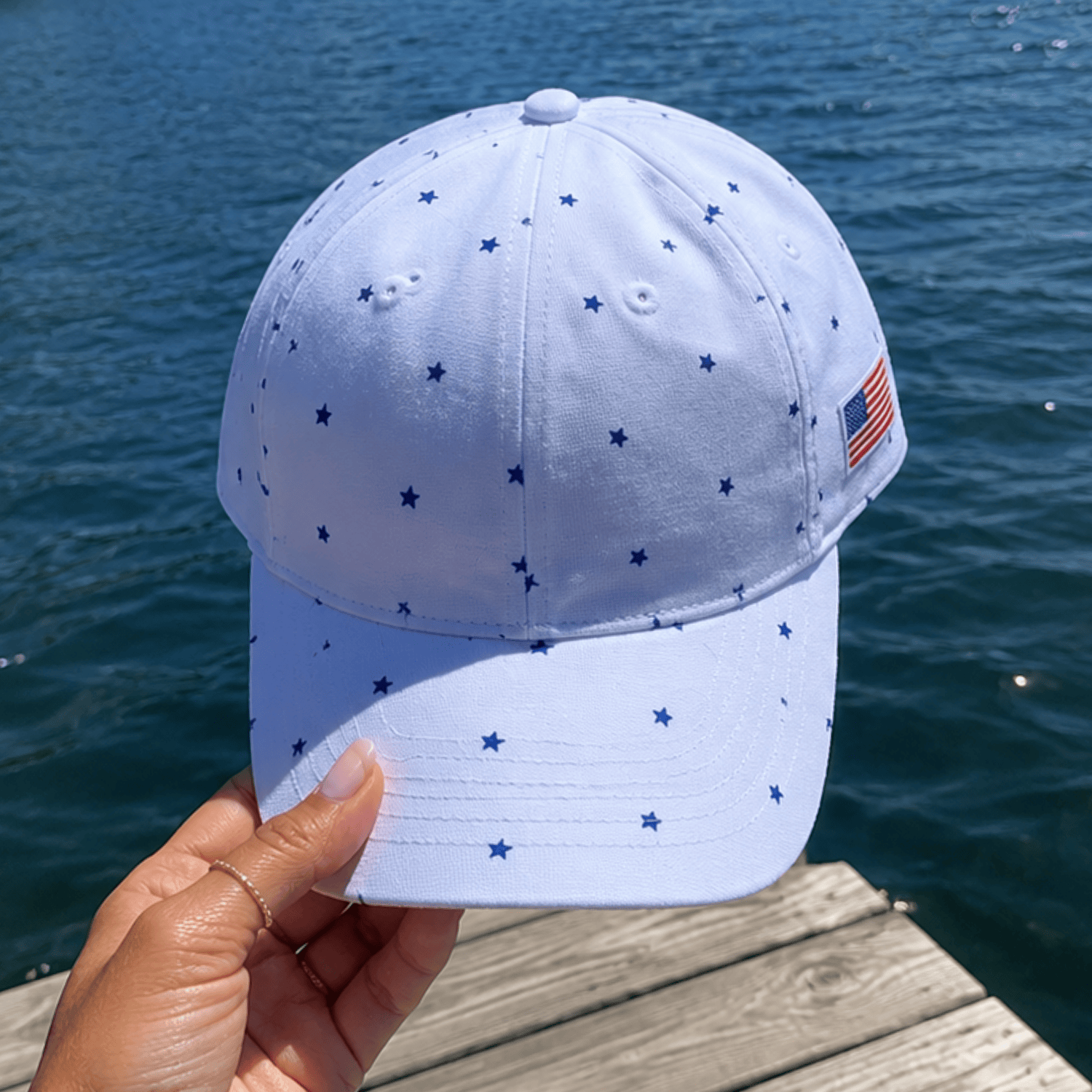 Starry Vibes Cap in light blue with navy stars and an American flag patch, held against a water background.